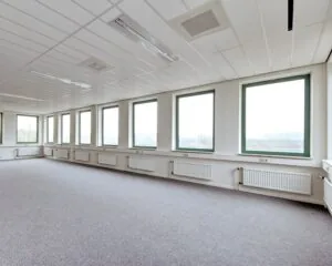 Lichte, lege kantoorruimte met groot raamoppervlak en grijze vloerbedekking.