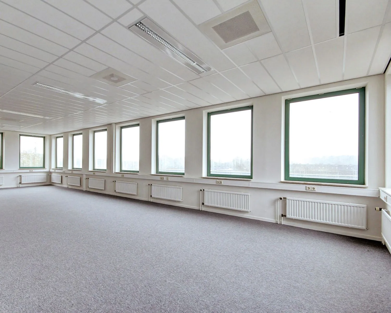 Lichte, lege kantoorruimte met groot raamoppervlak en grijze vloerbedekking.