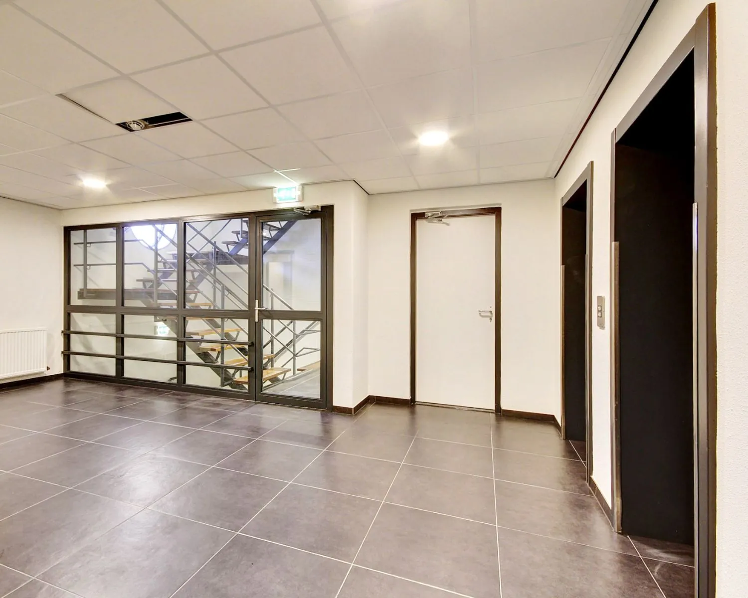Moderne, lege hal met twee liften, een trap achter glazen deuren en een gesloten witte deur.