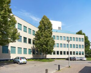 Modern kantoorgebouw met groen omrande ramen en geparkeerde auto's op een zonnige dag.