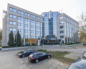 Moderne kantoorgebouw met spiegelende ramen en een parkeerplaats ervoor.