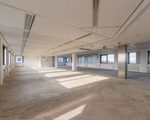 Lichte, lege kantoorruimte met grote ramen en een onafgewerkte betonnen vloer.
