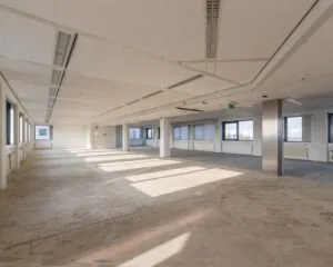 Lichte, lege kantoorruimte met grote ramen en een onafgewerkte betonnen vloer.