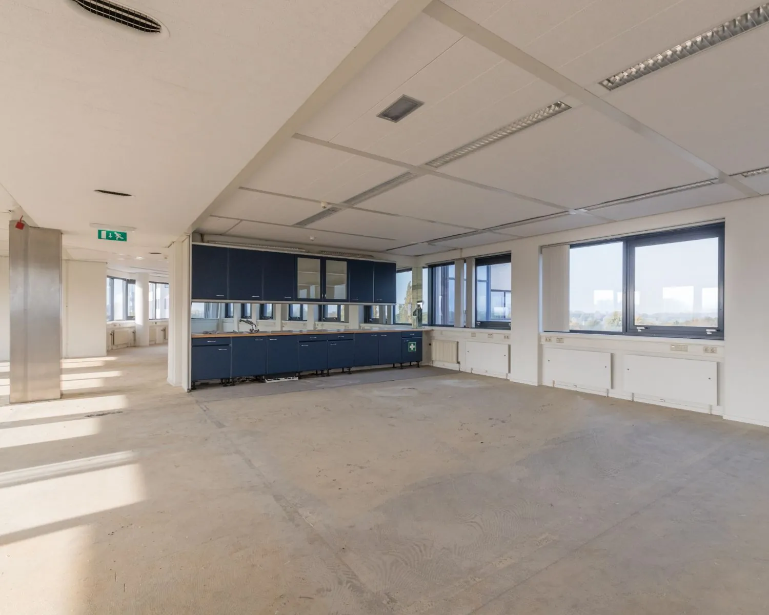 Lege kantoorruimte met blauwe ingebouwde keukenkastjes en grote ramen aan twee zijden.