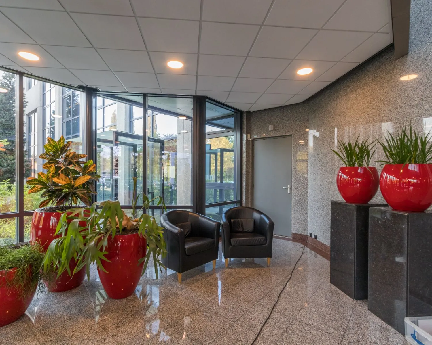 Moderne en lichte kantoorlobby met zwarte stoelen, grote rode plantenpotten en glazen buitenmuren.