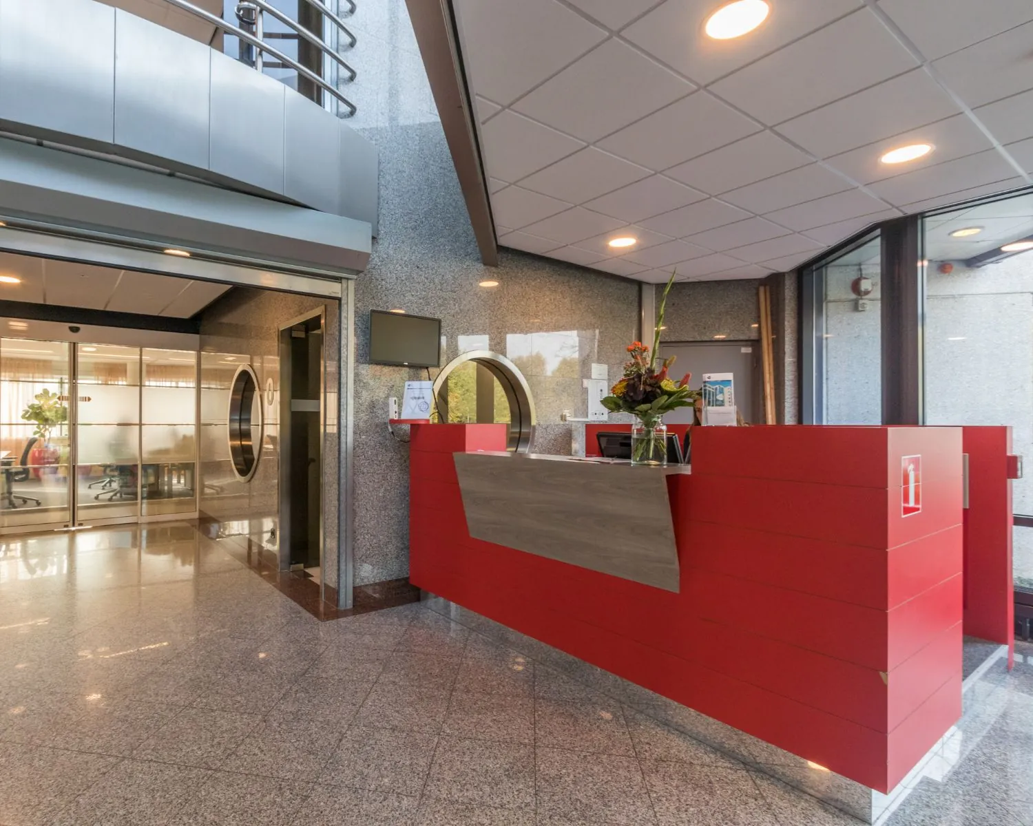 Moderne receptiebalie met rode panelen en een glazen bloemenvaas in een strak kantoorgebouwinterieur.