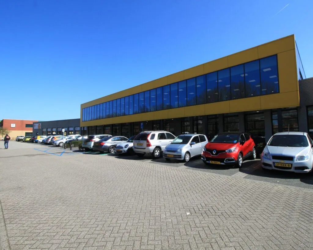 Parkeerplaats met geparkeerde auto's voor een modern bedrijfsgebouw met gele gevel en grote ramen.