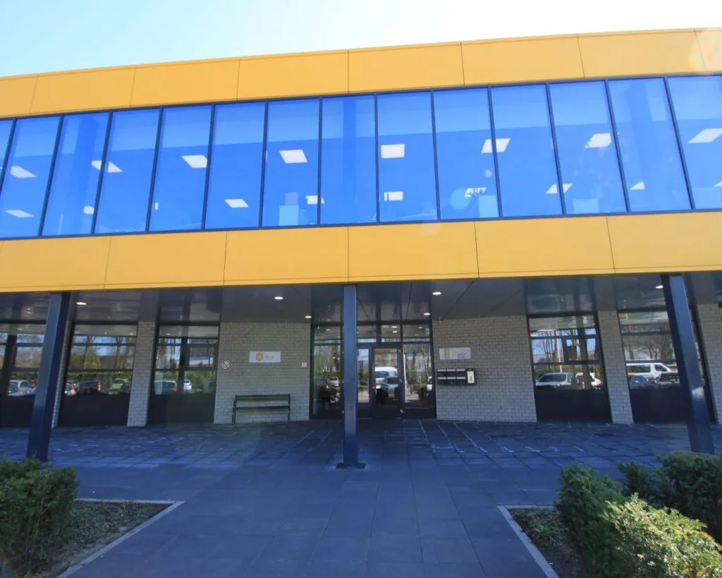 Vooraanzicht van een modern schoolgebouw met een gele gevel en grote blauwe ramen.