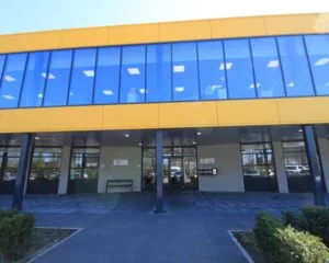 Vooraanzicht van een modern schoolgebouw met een gele gevel en grote blauwe ramen.