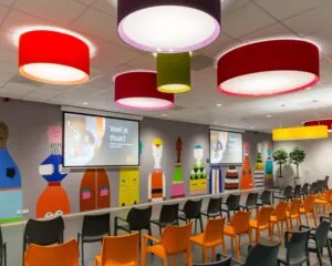 Kleurrijke presentatieruimte met felle plafondlampen, oranje en zwarte stoelen en een muurschildering van gestileerde figuren.