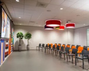 Kleurrijke presentatieruimte met opvallende hanglampen, oranje en zwarte stoelen, en een groot projectiescherm.