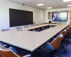 Moderne vergaderruimte met een U-vormige tafelopstelling, comfortabele stoelen en een presentatiescherm waarop "Voel je thuis!" staat.