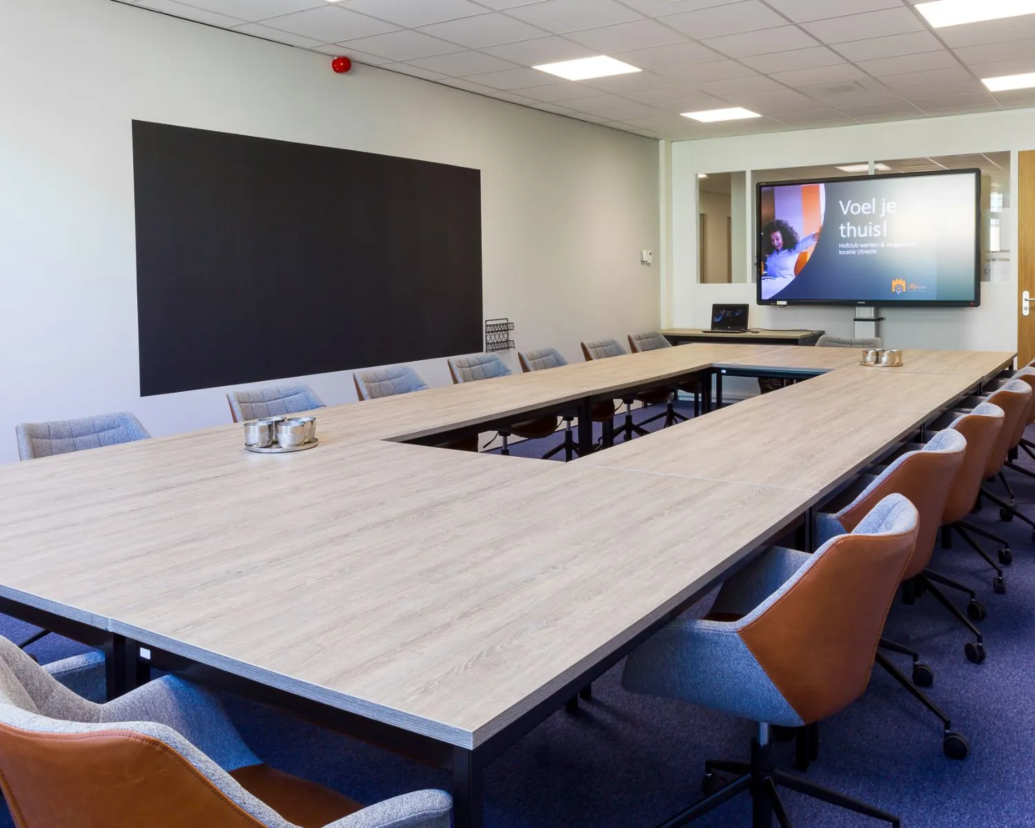 Moderne vergaderruimte met een U-vormige tafelopstelling, comfortabele stoelen en een presentatiescherm waarop "Voel je thuis!" staat.