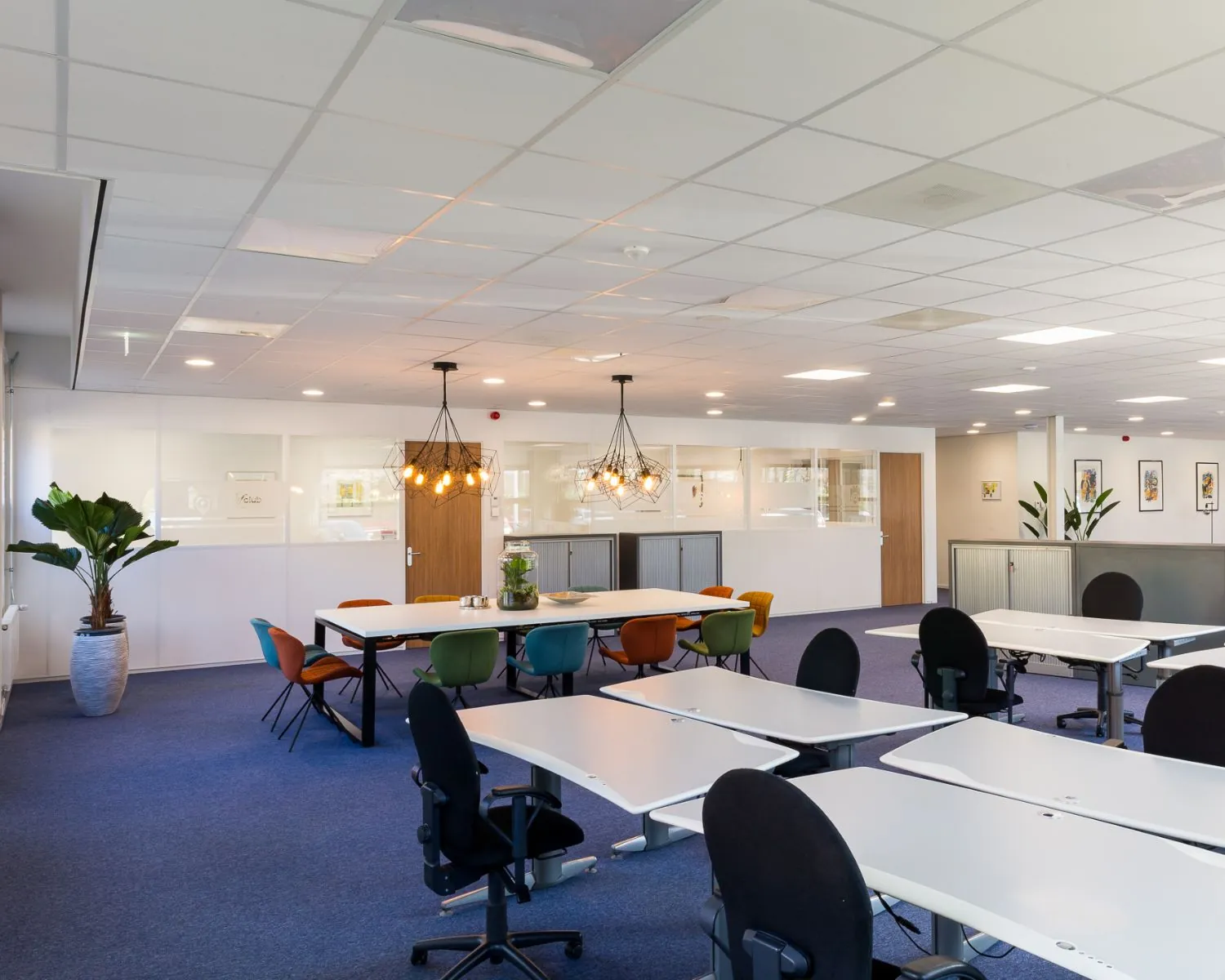 Moderne open kantoorruimte met flexwerkplekken, een vergadertafel met gekleurde stoelen en sfeerverlichting.