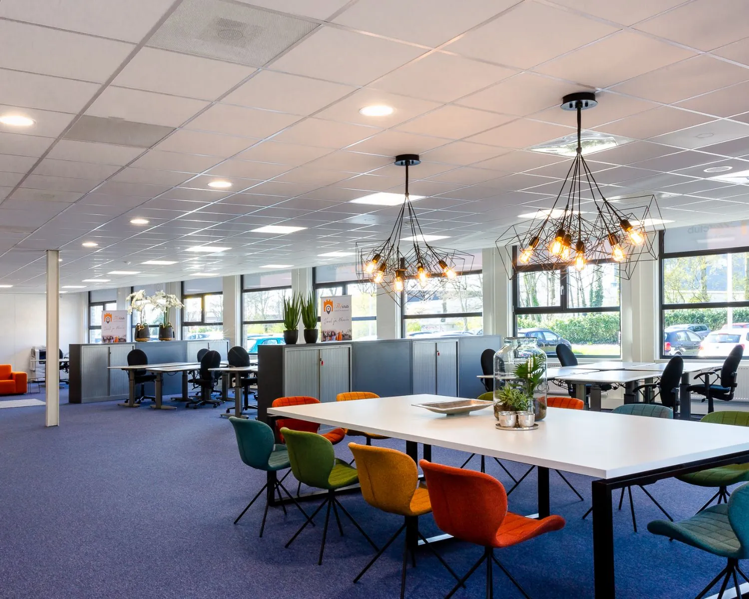 Moderne open kantoorruimte met kleurrijke stoelen, lange tafels en opvallende industriële hanglampen.