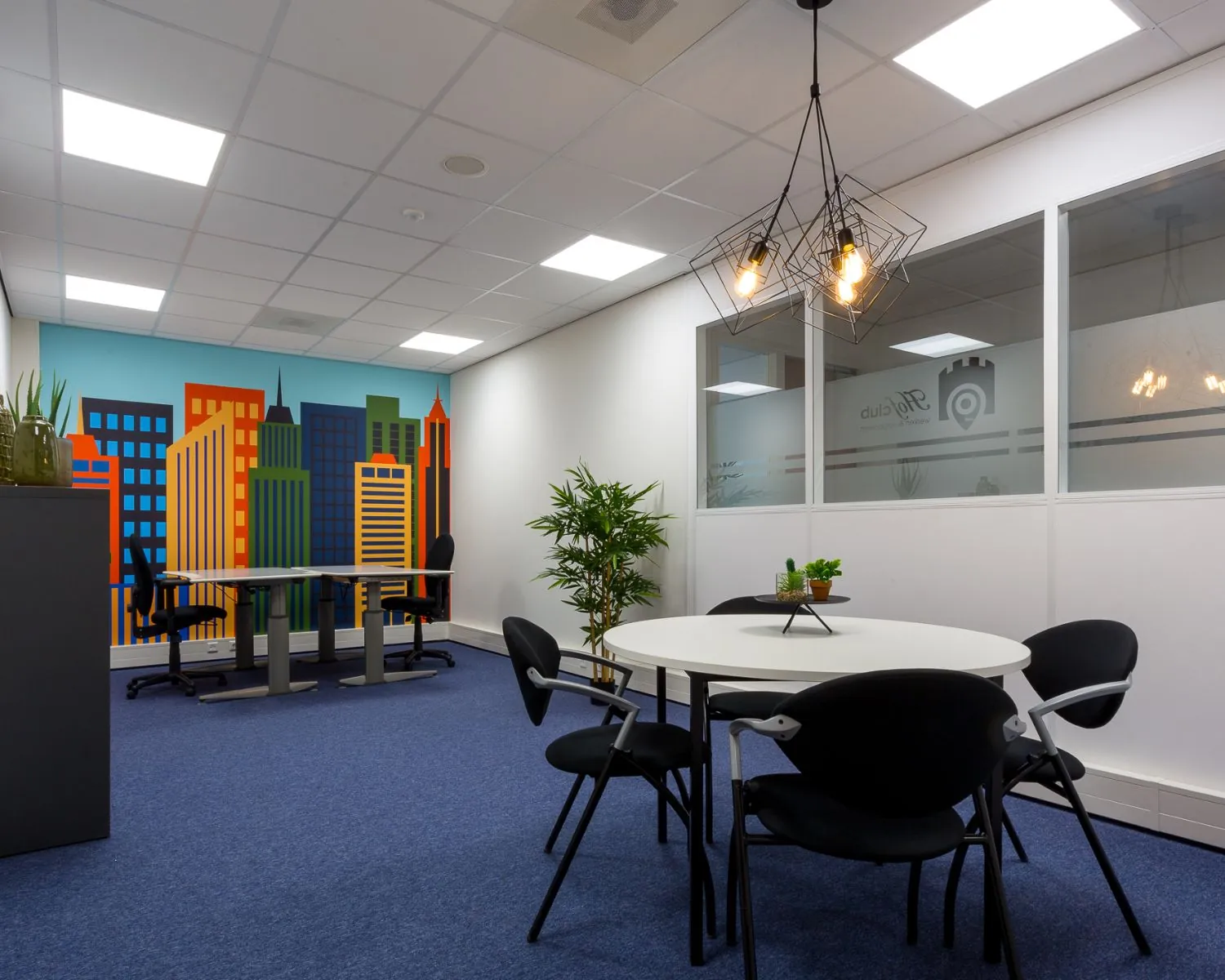 Moderne kantoorruimte met een ronde vergadertafel, kleurrijke muurschildering van een skyline en industriële hanglampen.