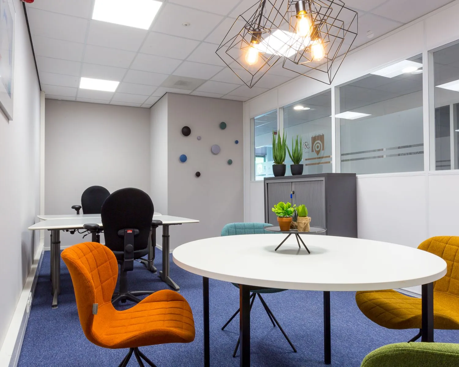 Moderne kantoorruimte met kleurrijke stoelen, ronde vergadertafel, hanglamp en planten op een kast.