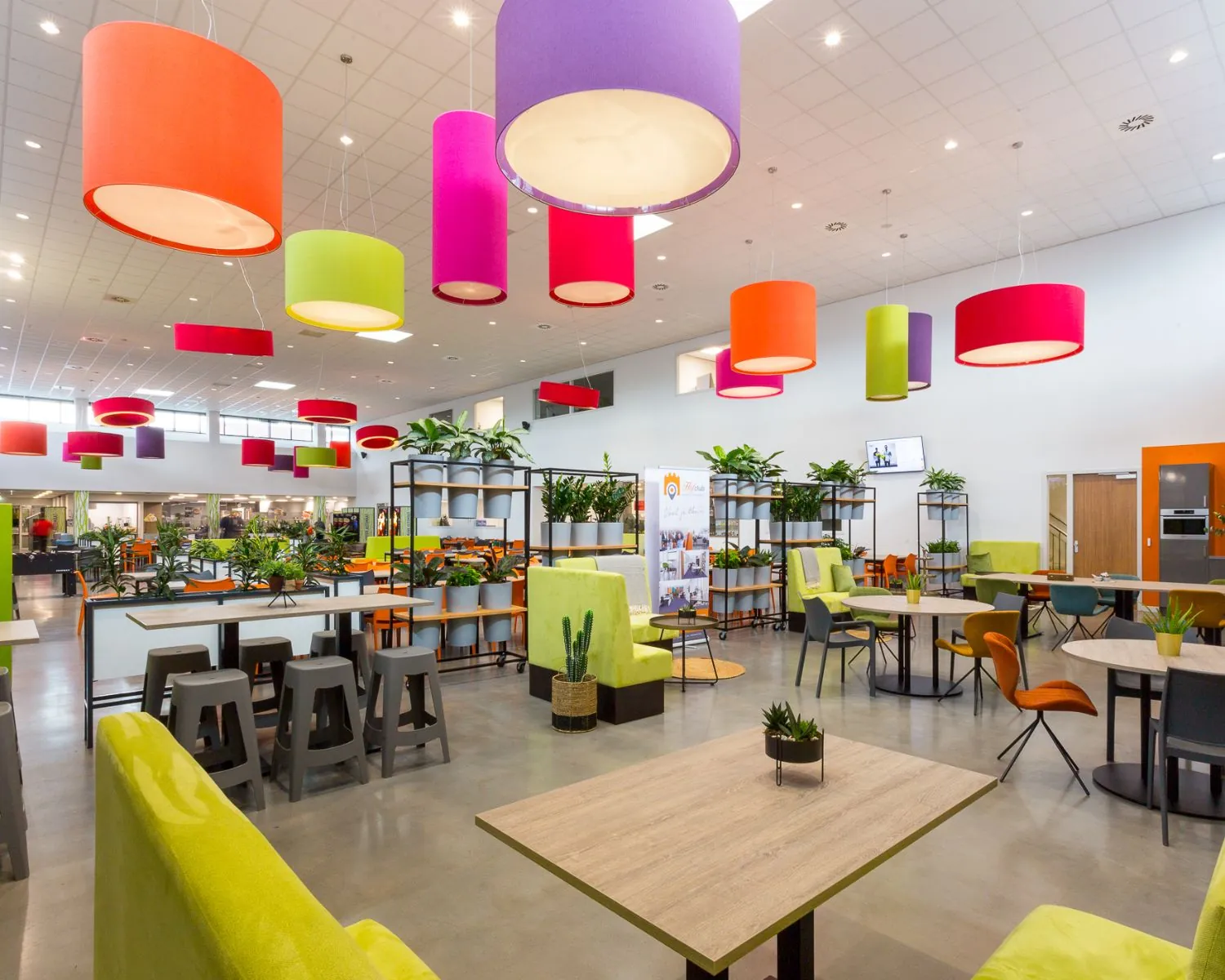 Moderne en kleurrijke kantine met felgekleurde hanglampen, groene planten en diverse zitmogelijkheden.