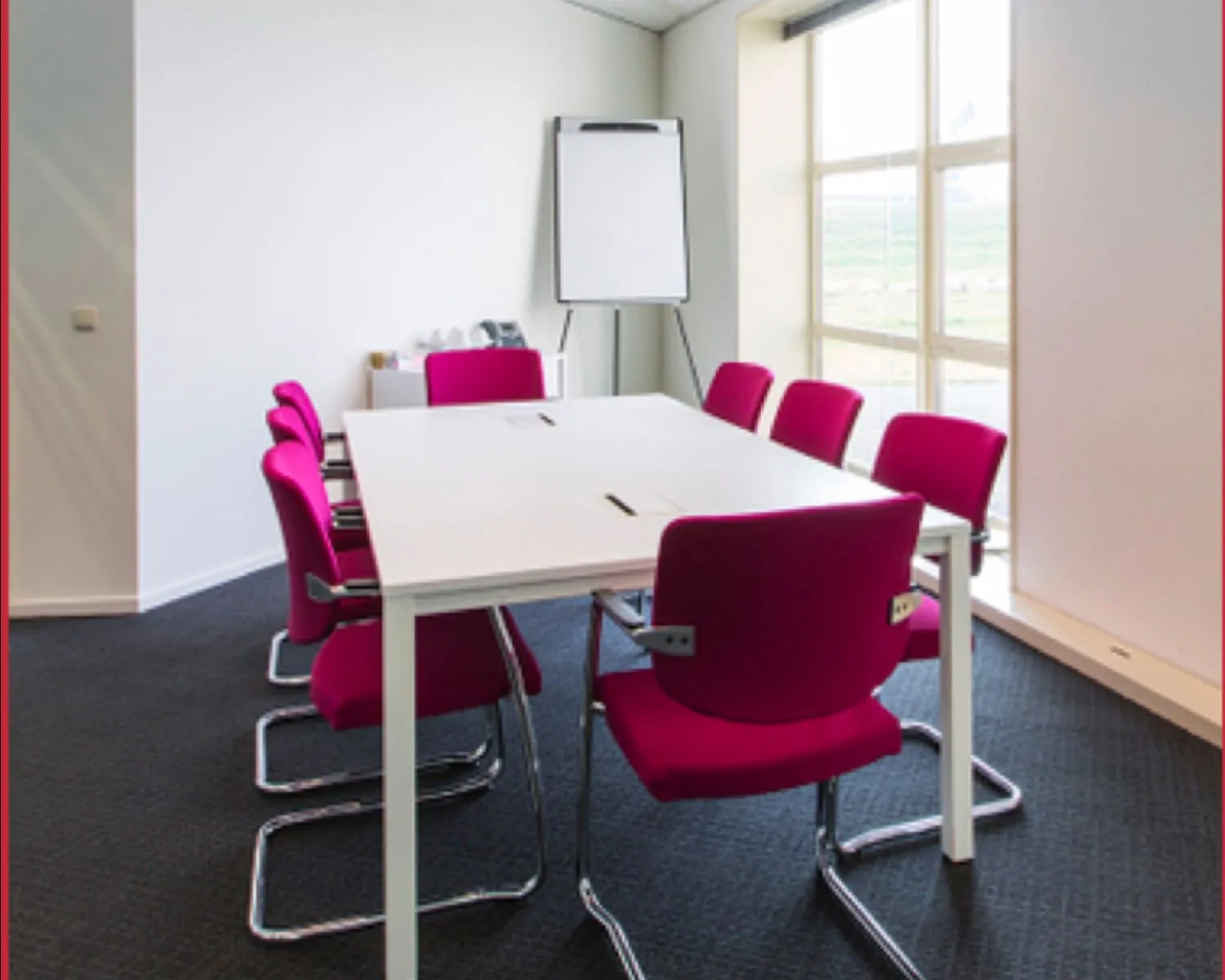 Vergaderruimte met een witte tafel, acht roze stoelen, een flip-over en grote ramen.