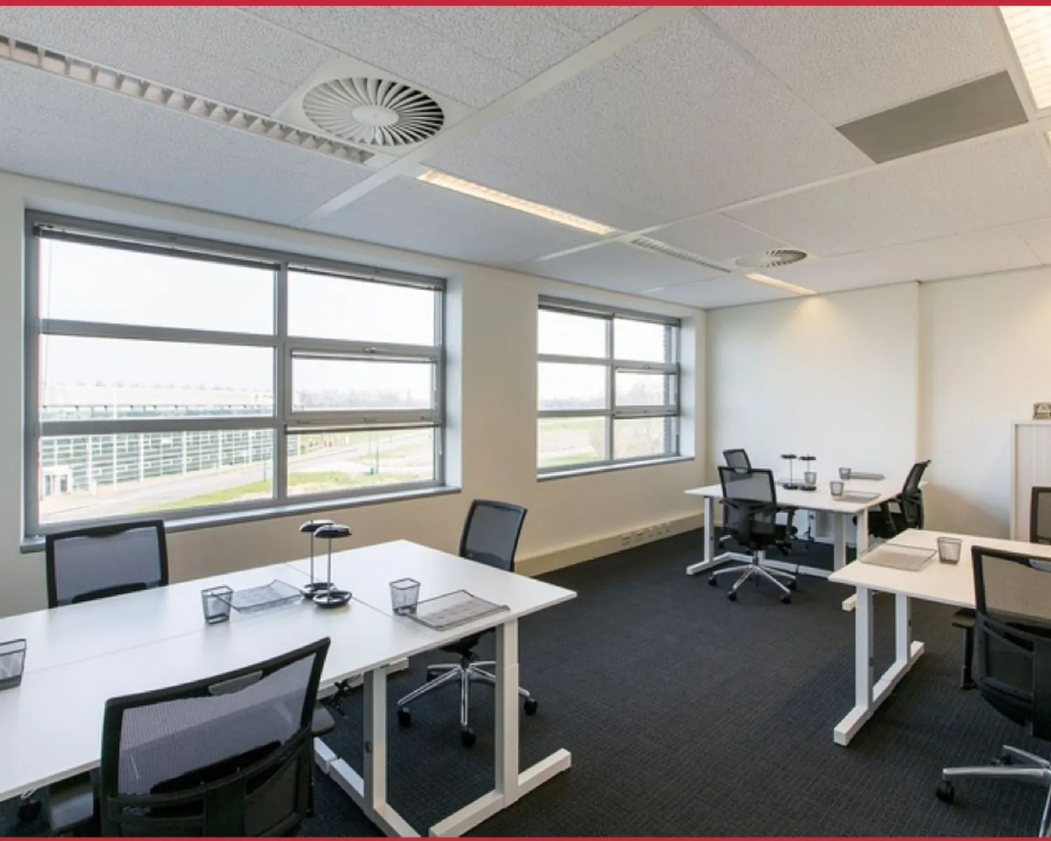 Modern, licht kantoor met vier werkplekken, grote ramen en ergonomische bureaustoelen.