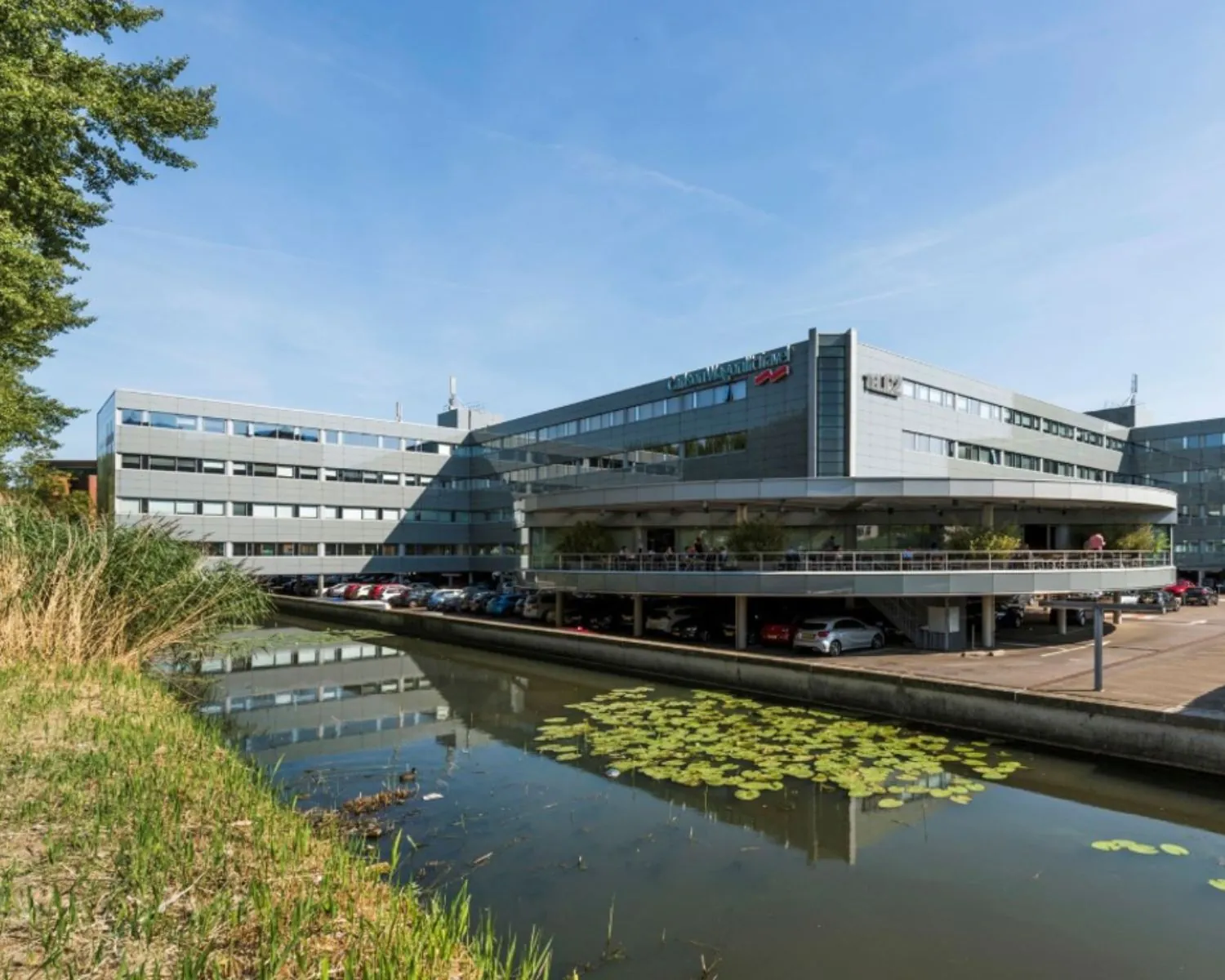 Kantoorgebouw "Croped_1557906213" met parkeergarage aan een vijver met waterlelies op een zonnige dag.