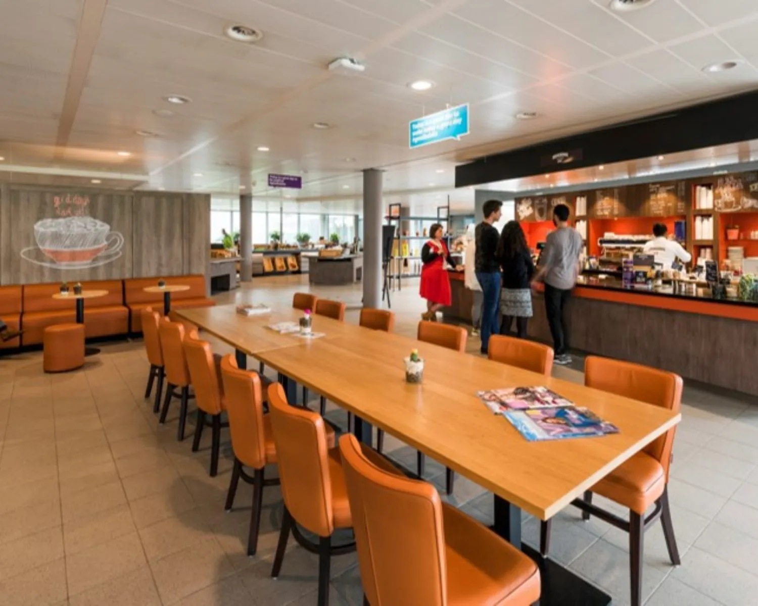 Moderne koffieruimte met oranje stoelen, een lange houten tafel en mensen bij de koffiebar.