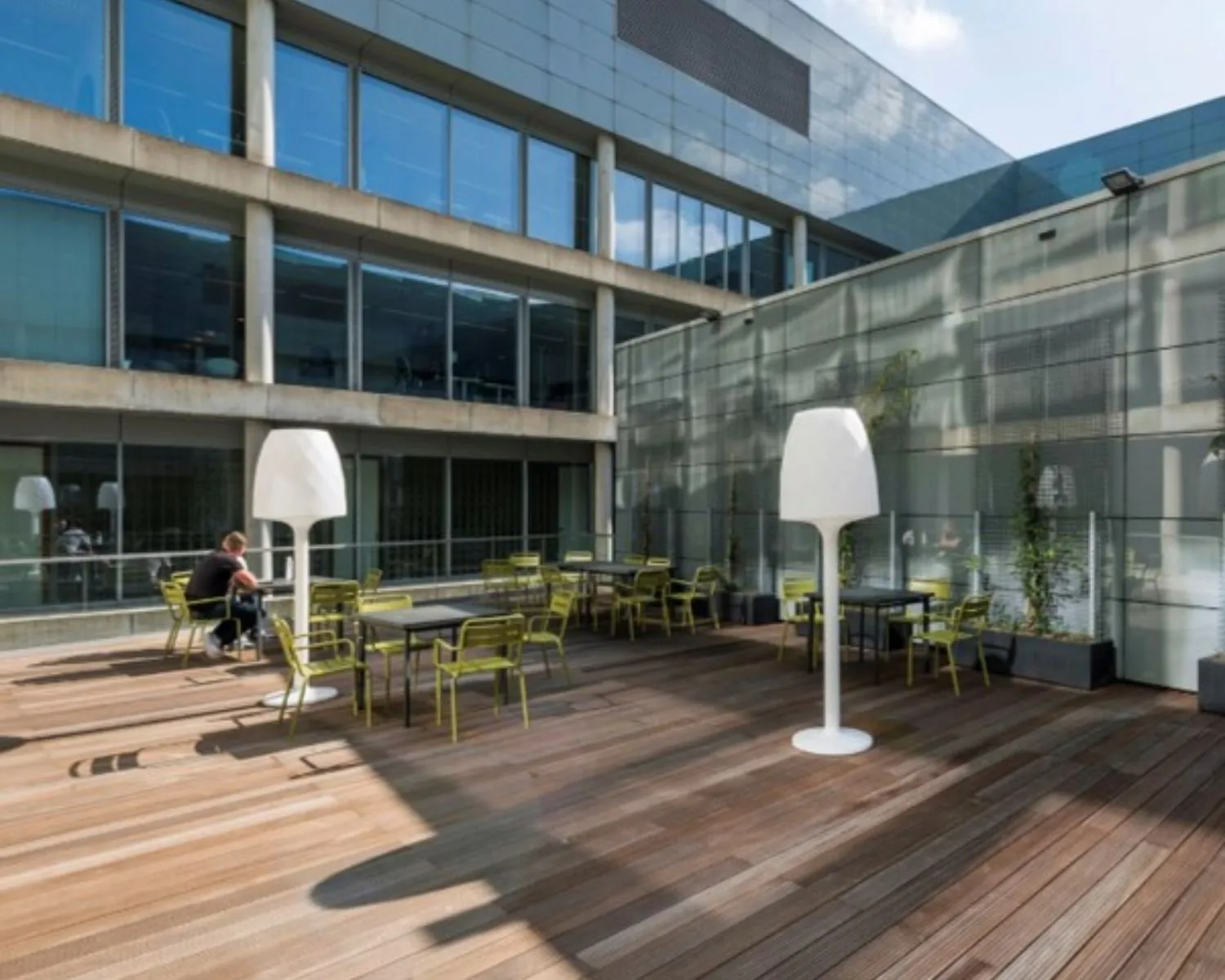 Moderne kantoorterras met houten vloer, groene stoelen, tafels en witte lampen, naast een glazen gevel.