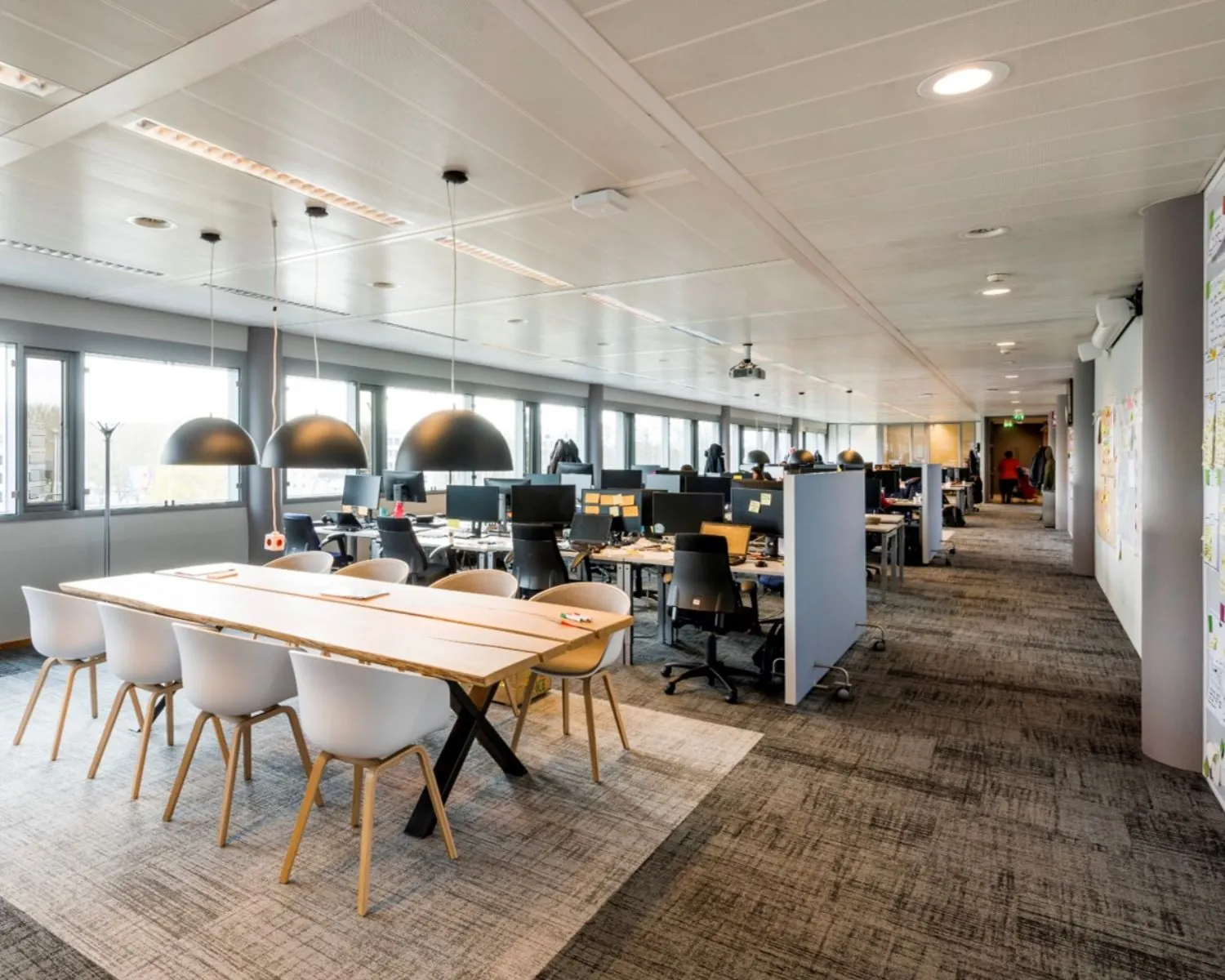 Moderne, open kantoortuin met bureaus, vergadertafel en grote ramen die veel daglicht binnenlaten.