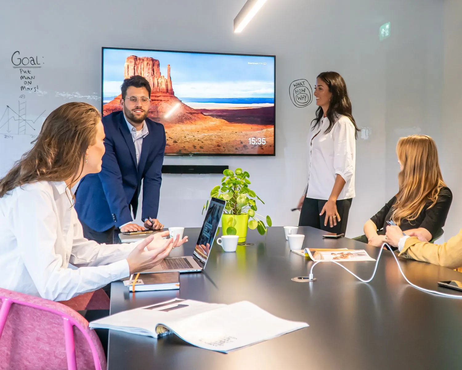 Vier mensen hebben een zakelijke vergadering in een modern kantoor met een groot scherm waarop een woestijnlandschap te zien is.