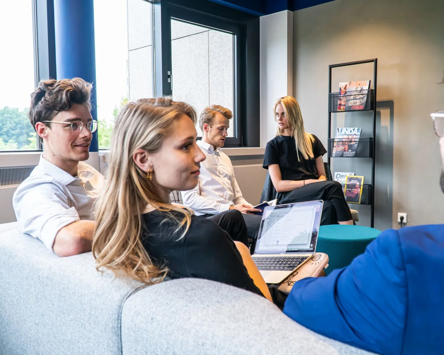 Een groep jonge professionals zit in een moderne kantoorruimte in overleg met een laptop en tijdschriftenrek op de achtergrond.
