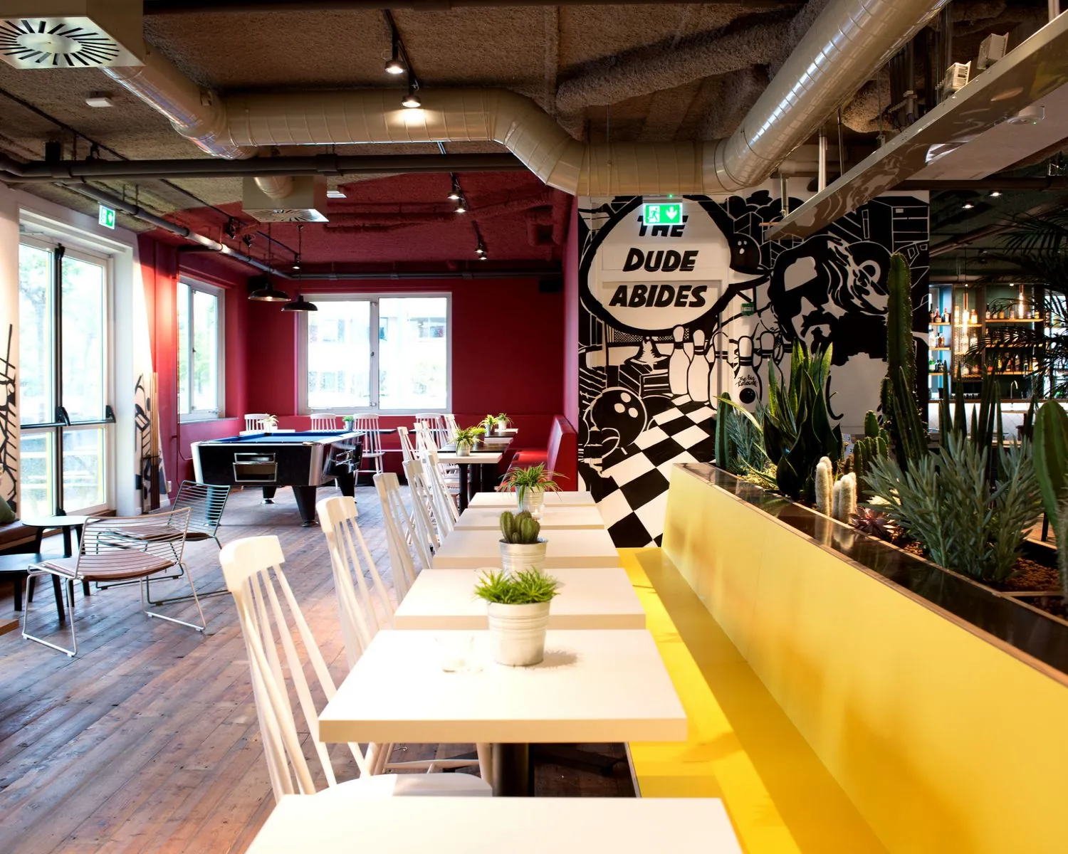 Modern ingerichte horeca-ruimte met gele bank, witte tafels, planten en een muurschildering met de tekst "The Dude Abides".
