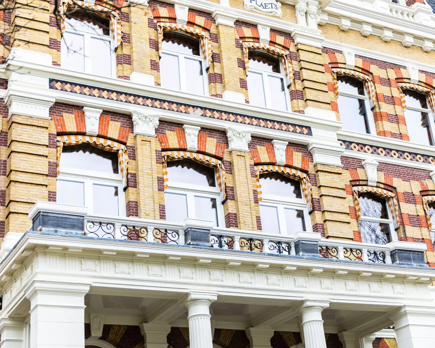 Detail van een historisch gebouw met decoratief metselwerk, witte ornamenten en smeedijzeren balkonrelingen.