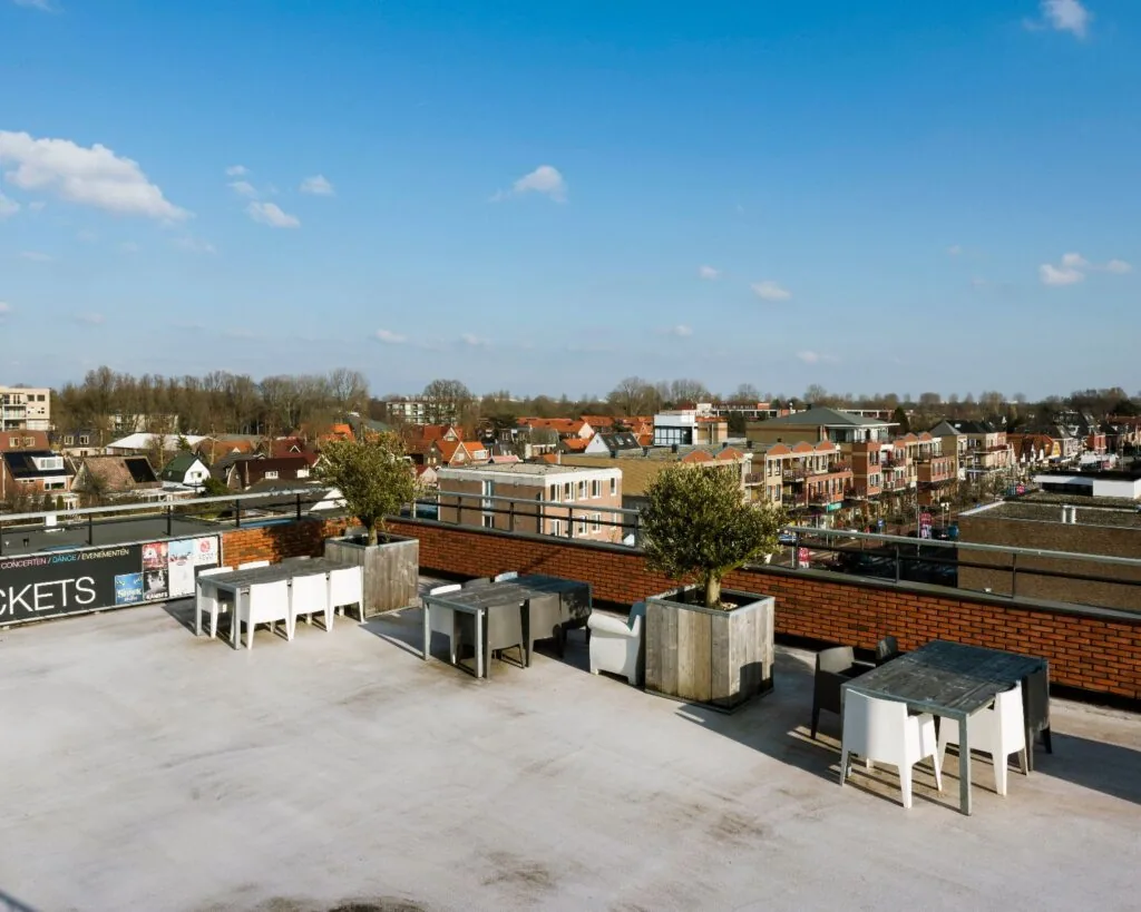 Zonnig dakterras met tafels en stoelen, uitzicht op een woonwijk met roodkleurige daken.