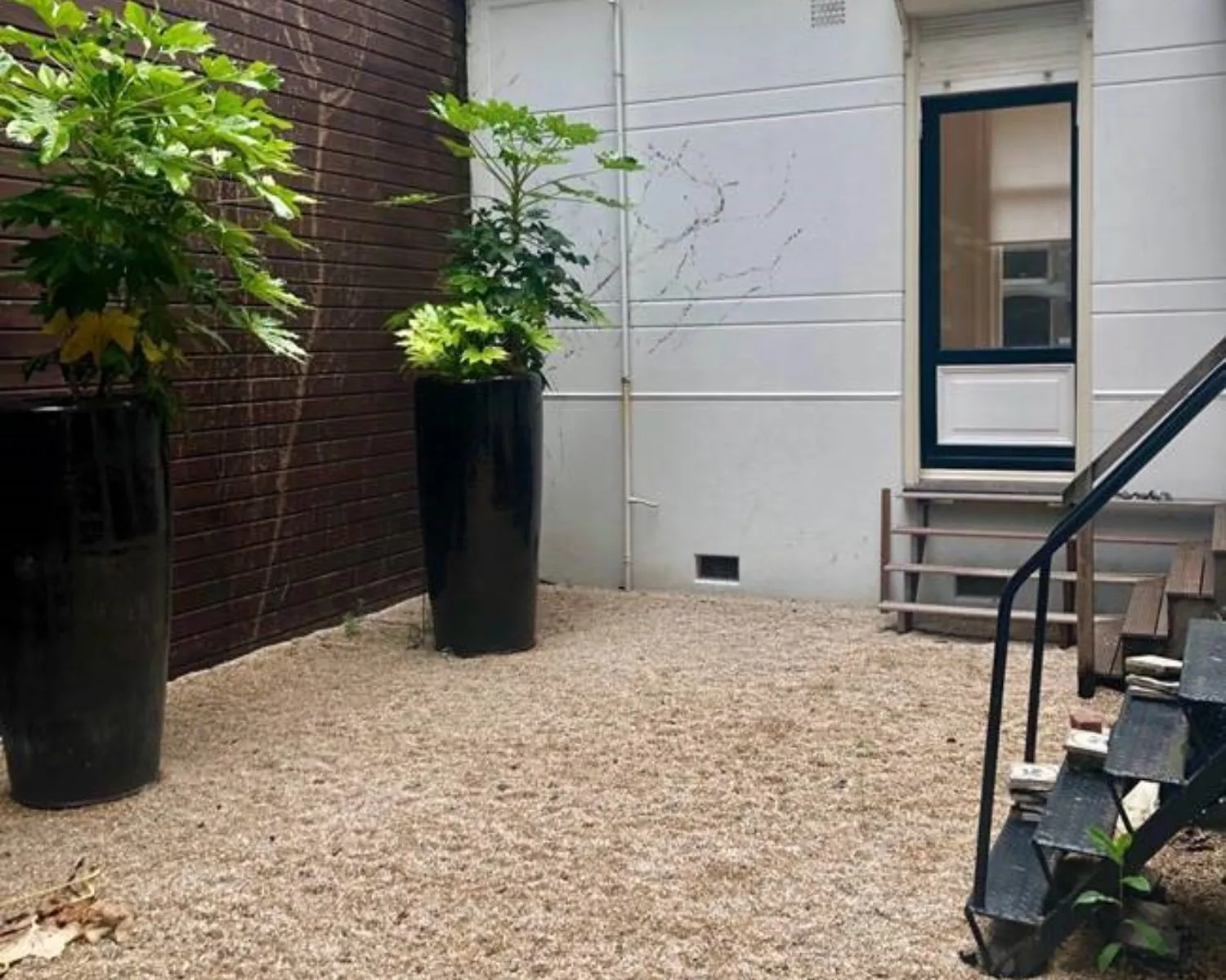 Kleine binnentuin met grind, twee hoge zwarte plantenpotten en een trap naar een witte achterdeur.