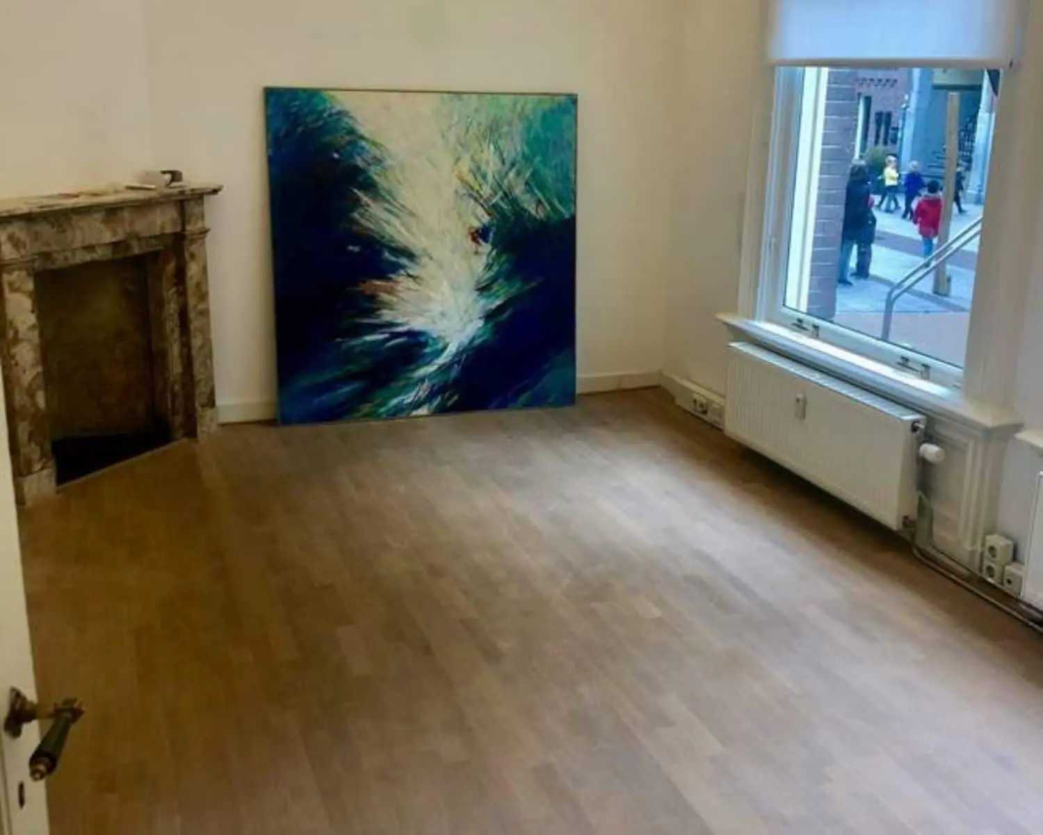 Lege kamer met houten vloer, een abstract schilderij tegen de muur en een raam met uitzicht op een winkelstraat.