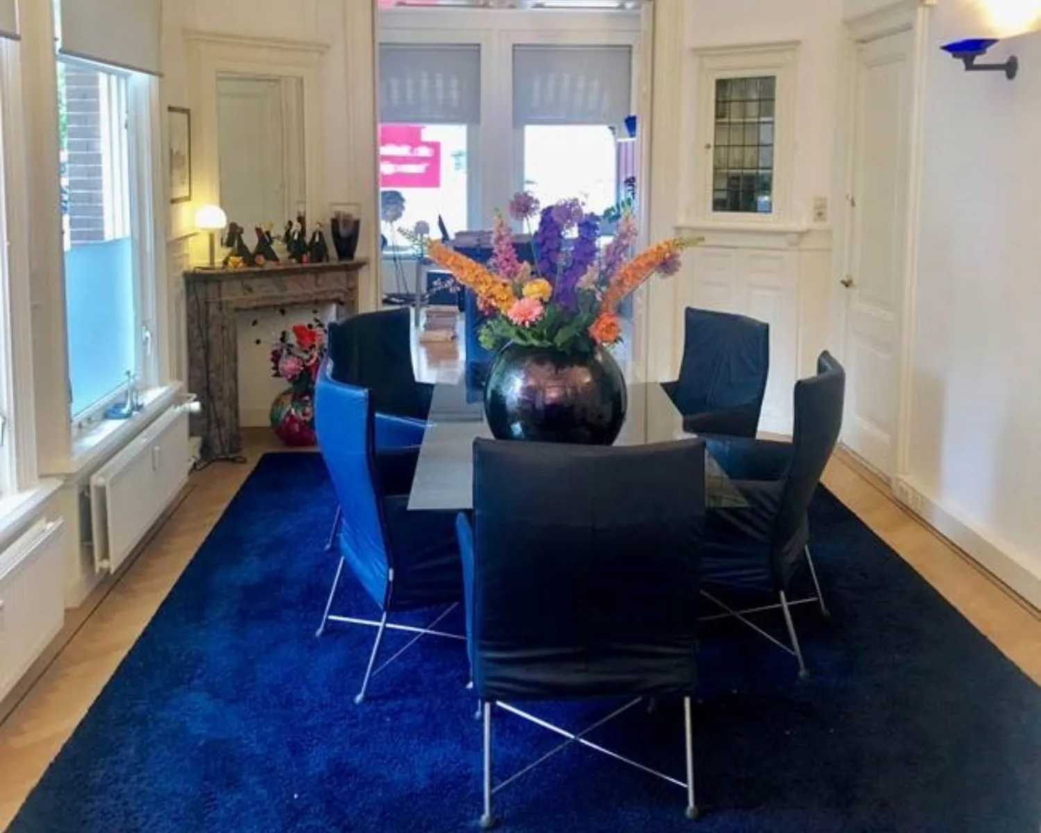 Vergaderruimte met zes blauwe stoelen rond een tafel, een grote vaas met kleurrijke bloemen en een donkerblauw vloerkleed.