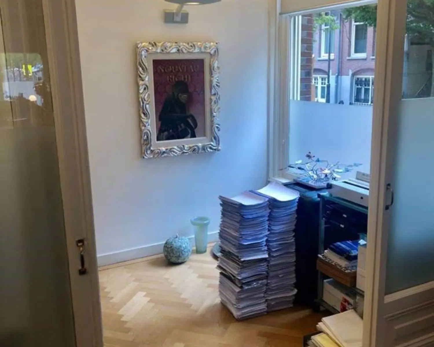 Kleine kantoorruimte met stapels papieren, een printer en een schilderij aan de muur.