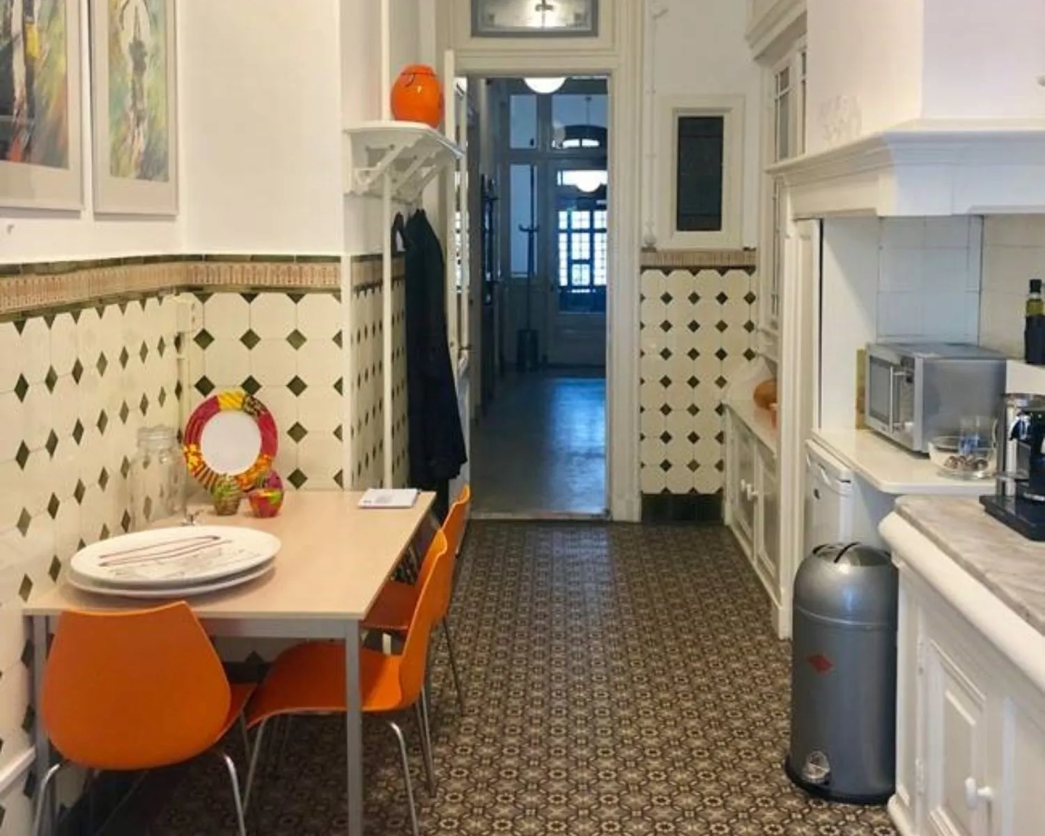 Keuken met retro tegelvloer, oranje stoelen en een smalle eettafel tegen de betegelde muur.