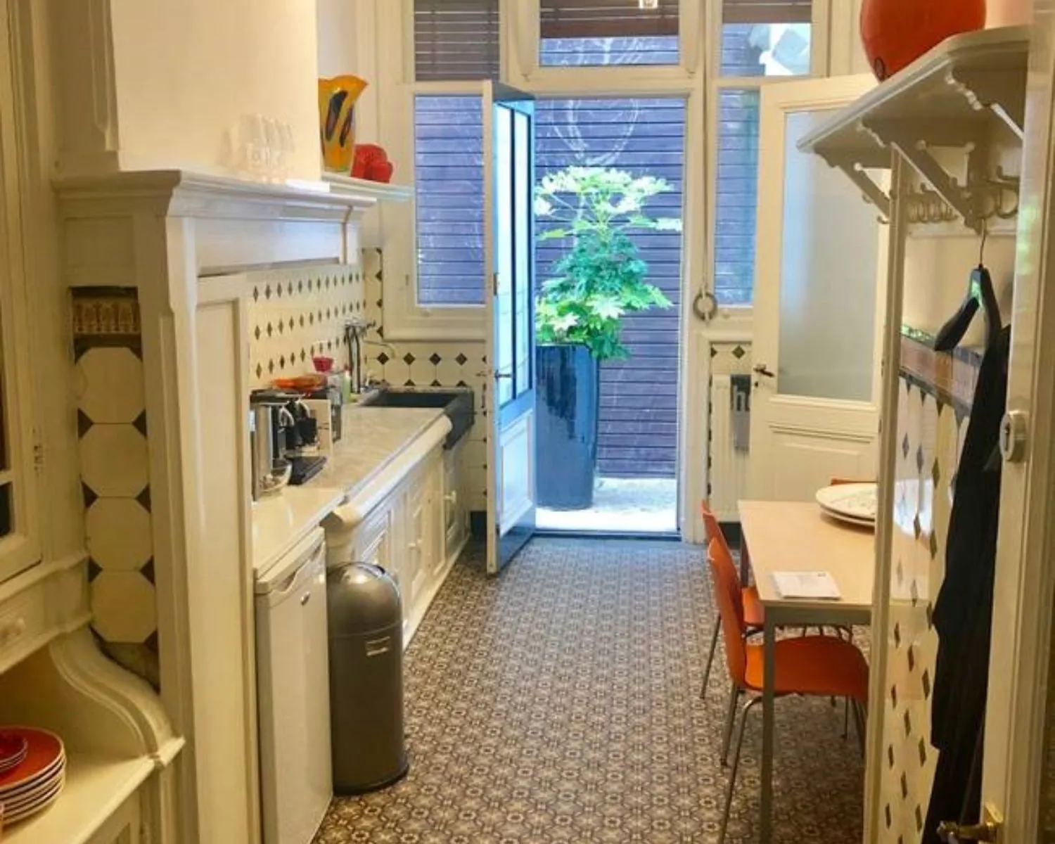 Lichte, smalle keuken met retrotegels, witte kasten, en een open deur naar een kleine patio met een grote plant.