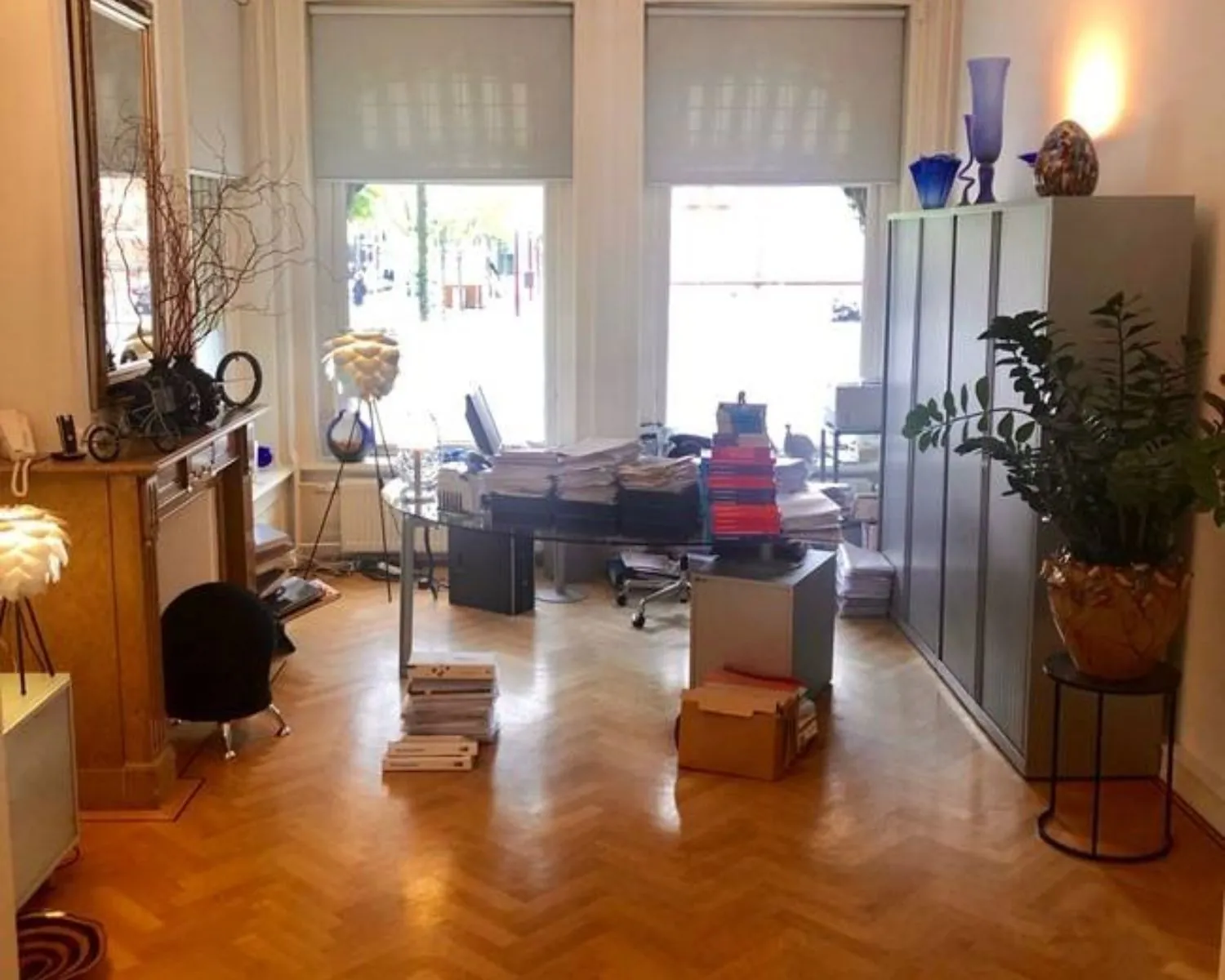 Een kantoorruimte met houten vloer, grote ramen, stapels dossiers en moderne decoratie.