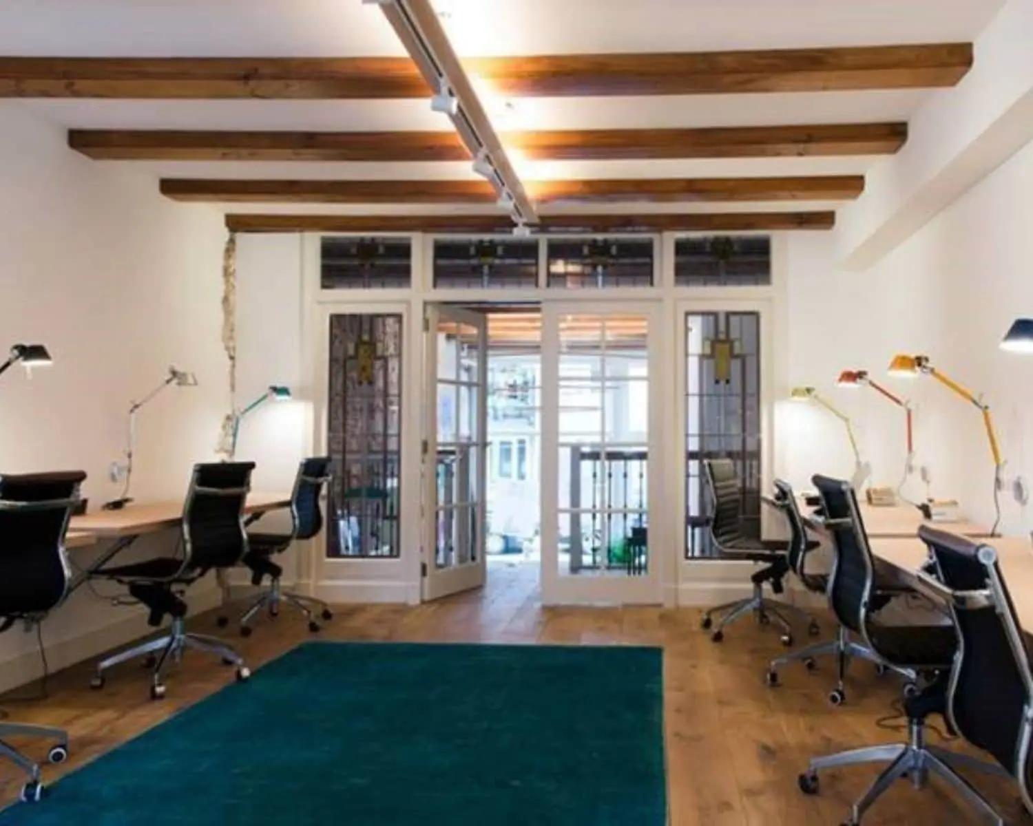 Modern ingerichte kantoorruimte met bureaus, bureaustoelen en gekleurde bureaulampen, met houten balken aan het plafond en glazen deuren aan het einde van de ruimte.