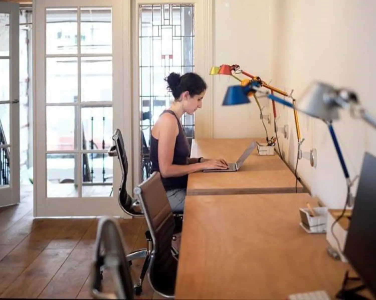 Vrouw werkt op een laptop aan een lange houten bureautafel in een rustige, moderne werkruimte met bureaulampen.