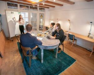 Vier mensen hebben een vergadering in een sfeervolle kantoorruimte terwijl een vrouw iets uitlegt op een whiteboard.