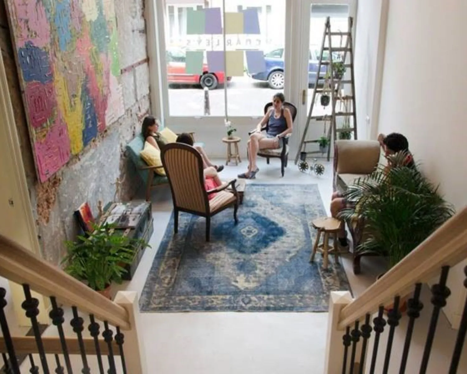 Vier mensen zitten ontspannen in een gezellig ingerichte woonkamer met vintage meubilair, planten en kleurrijke kunst aan de muur.