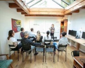 Een groep mensen volgt een presentatie van een vrouw in een lichte vergaderruimte met glazen dak.
