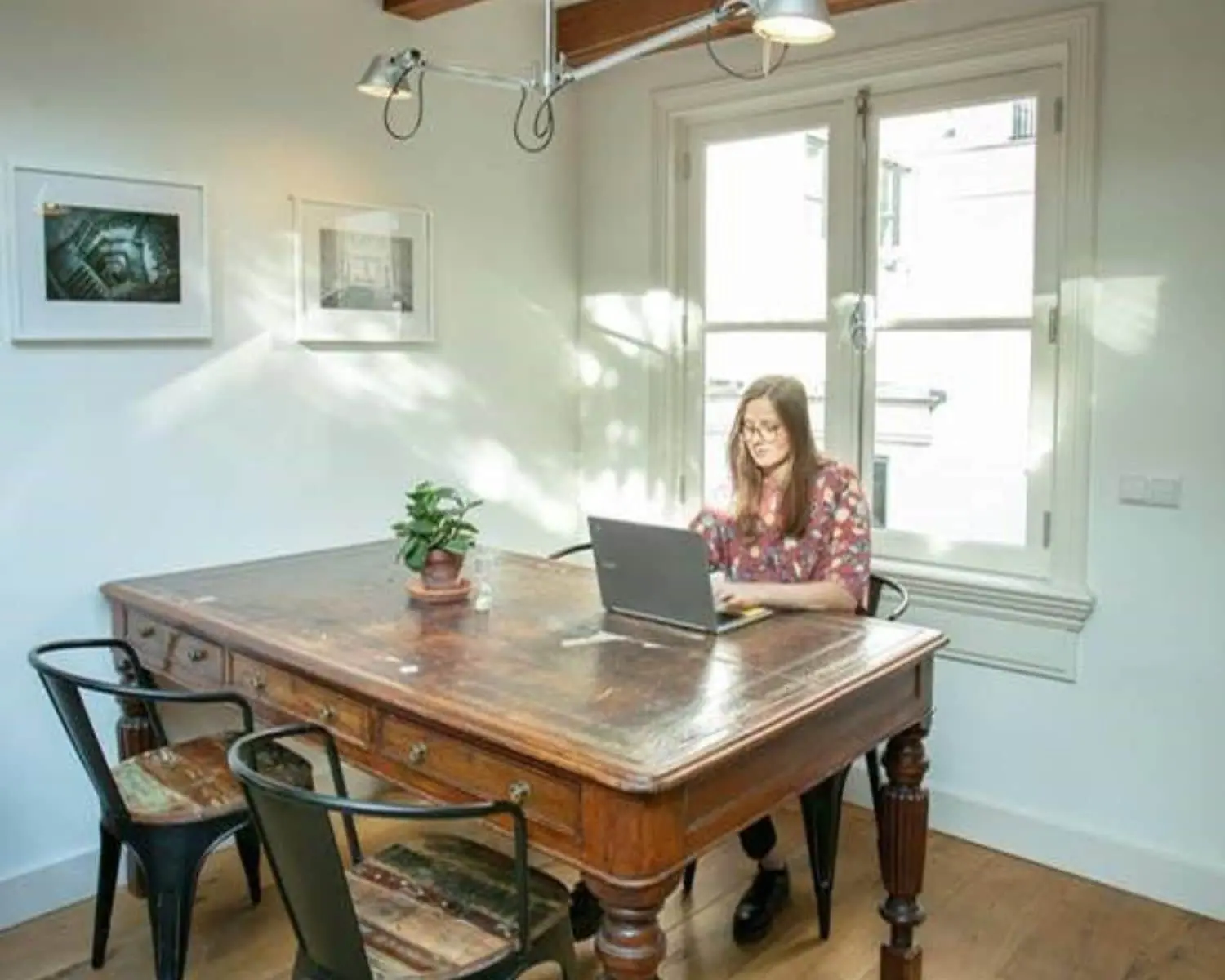 Vrouw werkt op een laptop aan een grote houten tafel in een licht kantoor met kunst aan de muur.