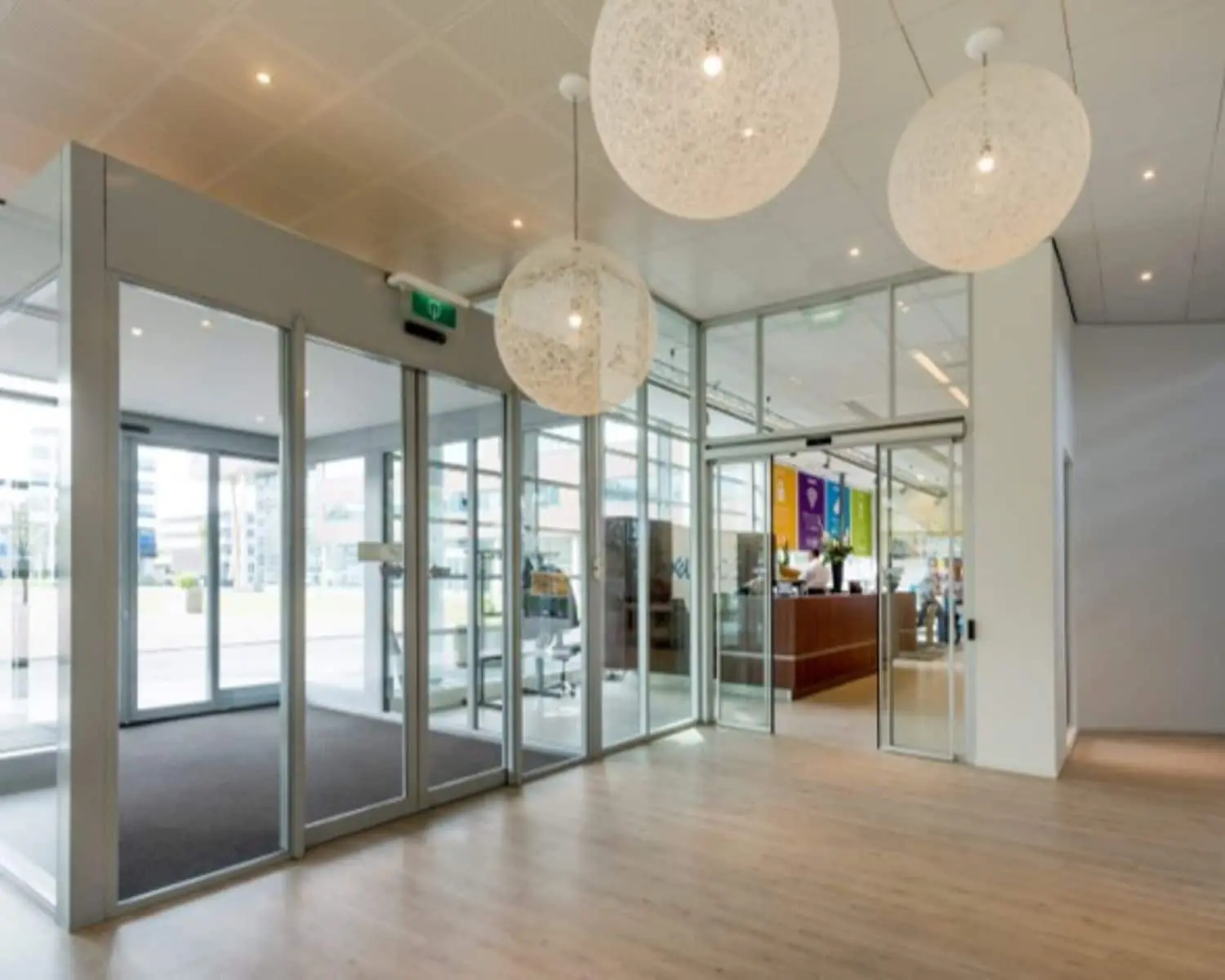 Moderne kantoorentree met glazen schuifdeuren, houten vloer en grote ronde hanglampen.