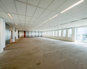 Leegstaand kantoor met systeemplafond, onafgewerkte vloer en grote ramen langs de rechterzijde.