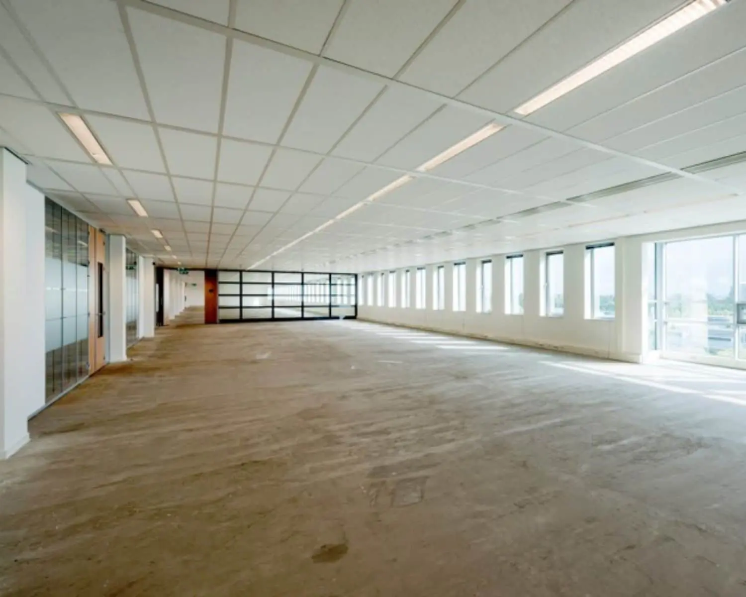 Leegstaand kantoor met systeemplafond, onafgewerkte vloer en grote ramen langs de rechterzijde.