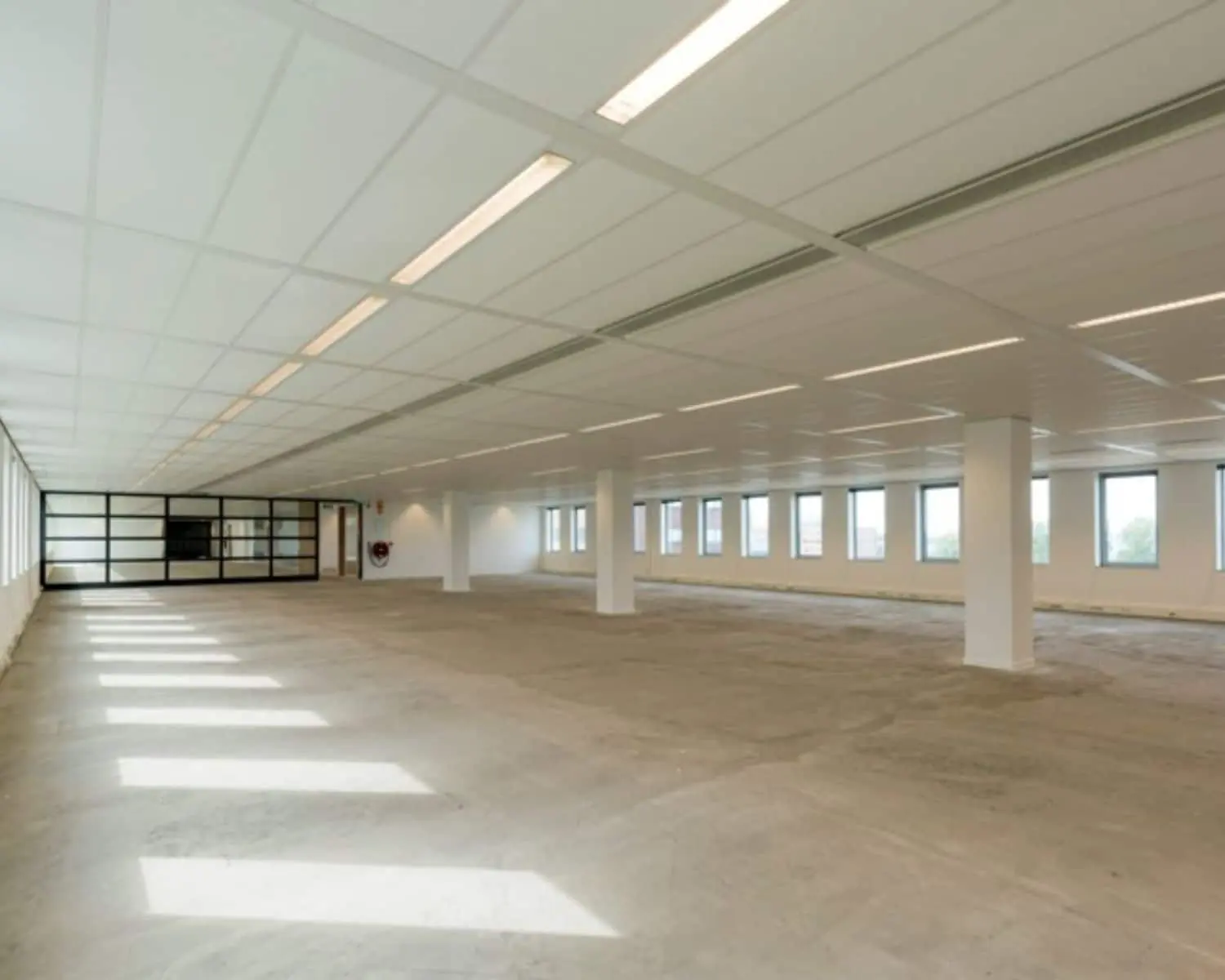 Leeg, modern kantoor met systeemplafond, betonnen vloer en grote ramen aan beide zijden.